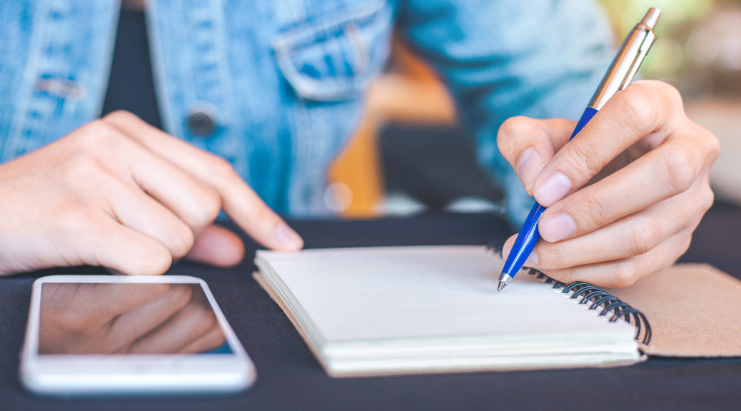 Woman hand is writing on a note pad with a pen and the phone is placed on the table